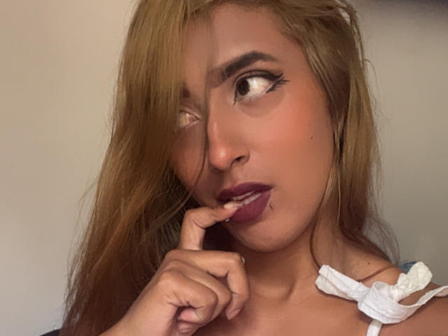 SophiaForeest mit langem, blondem Haar, braunen Augen und lila Lippenstift, die ihren Finger beißend mit einem schmollenden Gesichtsausdruck, trägt ein weißes Oberteil mit gerüschten Trägern..
