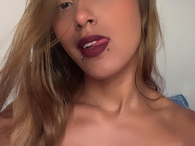 SophiaForeest heeft een bruine huid, lang blond haar, donkerrode lippenstift, een lip-piercing en haar tong steekt een beetje uit. Ze draagt geen top en haar sleutelbeenderen zijn zichtbaar..