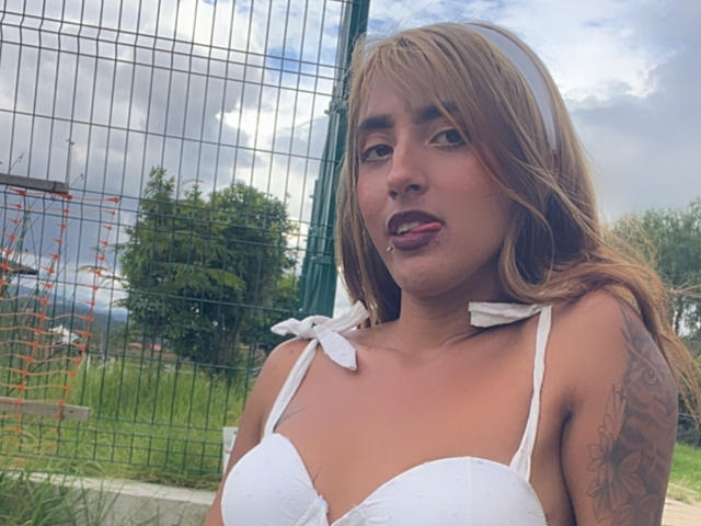 SophiaForeest met een bruine huid, blond haar, een witte beha, haar tong uitgestoken, een tatoeage op haar rechterarm, een witte hoofdband, staand voor een groen metalen hek, bewolkte lucht. Borsten, beha, tong..