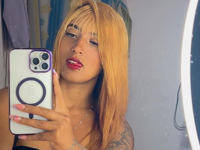 SophiaForeest with blonde hair, red lipstick, and tattoos, holding an iPhone, taking a mirror selfie with her finger on her glossy pink nail. Her tongue is slightly out.