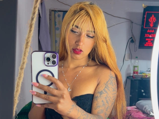 SophiaForeest, met lang blond haar, rode lippenstift en tatoeages op haar arm, houdt een telefoon vast met een ringhouder, draagt een zwarte top zonder bandjes en maakt een selfie in de spiegel..