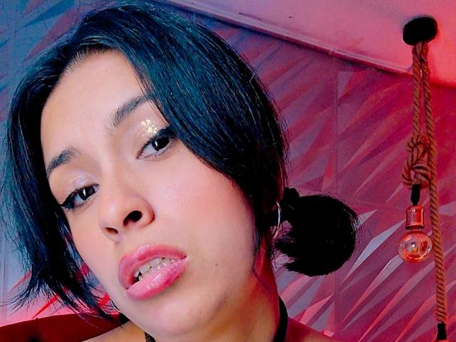 RoxanneHan with black hair in puffs, shiny pink lips, glittery eyeshadow, and a slight pout, wearing a black choker, has a rope and light bulb in the background.