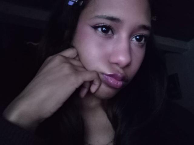 LovelyConnie has brown skin, dark wavy hair, and is touching her cheek with her right hand. She has purple lipstick and winged eyeliner, looking slightly sad.