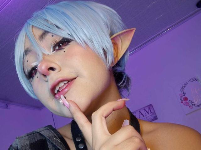 IvonnySakura com cabelo curto azul, orelhas de elfo e maquiagem, dedo perto dos lábios rosados, top de alça preta, luz roxa, unhas compridas cor de rosa, sorrindo, fofa, visual de fantasia..