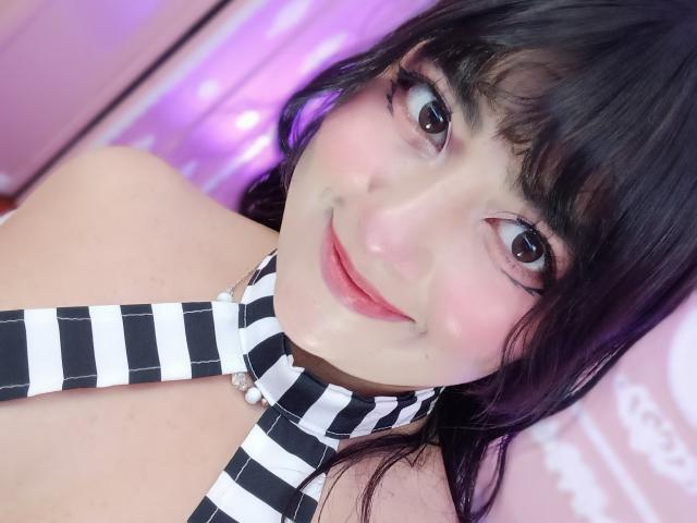 NekoPam has black striped necktie, pale skin, and dark wavy hair with bangs, smiling with pink lips, brown eyes, and subtle blush, in a pink-lit room.