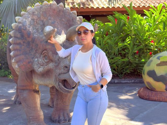 KattyRivera with big tits, white shirt, blue jeans, pink cardigan, white cap, sunglasses, hand in pocket, touching dinosaur statue's nose. Dinosaur has textured brown skin, green plant background.