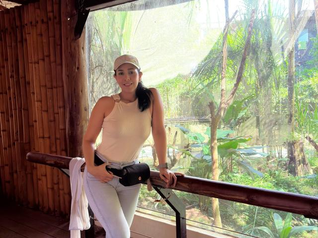 KattyRivera with black ponytail, white cap, sleeveless top, white pants, leaning on wooden railing, holding camera, white shirt draped over railing, green jungle outside, sunny, smiling.