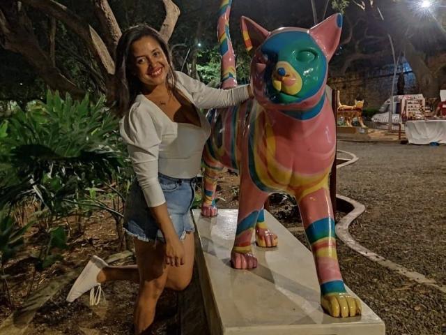 MirandaDreams with a colorful, painted cat statue. She's wearing a white jacket, brown top, and denim shorts, standing next to the cat with her hand on it. Nighttime.