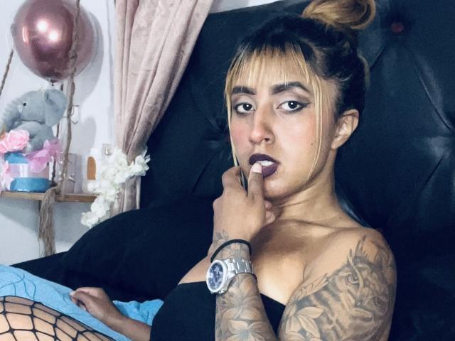 SophiaForeest, com os seios à mostra, usando meias de rede, tem tatuagens no braço e está chupando o dedo enquanto está sentada em uma cama preta. Seu cabelo está preso em um coque..