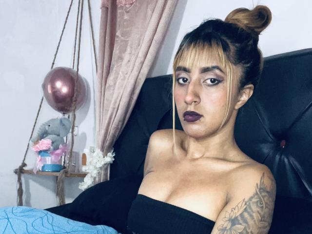 SophiaForeest, avec une peau mate, des cheveux bruns attachés en chignon, du rouge à lèvres foncé, un bras tatoué, un haut noir sans bretelles, assise sur un lit noir, avec un rideau rose, un ballon rose, un éléphant en peluche et des fleurs..