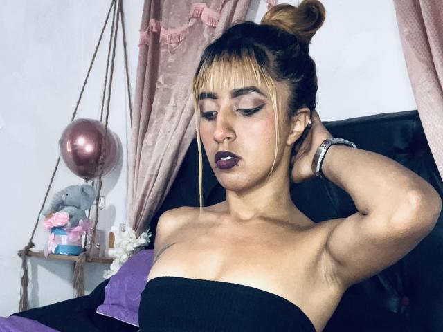 SophiaForeest, con il seno scoperto, braccia alzate, rossetto scuro, capelli biondi raccolti in uno chignon, tende rosa, vestito nero con spalline, cuscino viola, palloncino rosa e un elefante di peluche sullo sfondo..