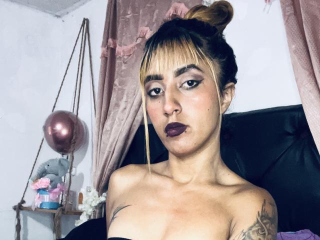 SophiaForeest met een blonde knot, donkere lipstick, tatoeages en ontblote borsten, kijkt in de camera met volgeblazen lippen, met roze gordijnen en een roze ballon op de achtergrond..