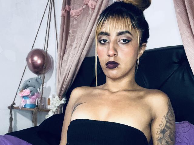 SophiaForeest, con lápiz labial oscuro, flequillo rubio y un top negro sin tirantes, mostrando sus pechos y su brazo tatuado, sentada en una cama negra, mirando a la cámara..