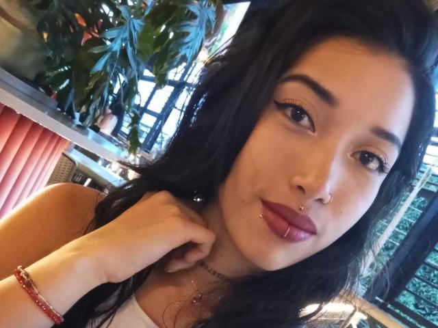 KrystalEvans with dark hair, glossy pink lips, nose ring, and eyeliner, holding her neck, wearing a white top, pink bracelet, and necklace, in a sunlit, plant-filled setting.