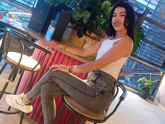 KrystalEvans with dark hair, white tank top, tight grey jeans, and white sneakers, sitting on a chair, smiling, showing her ass, and a tattoo on her left arm.