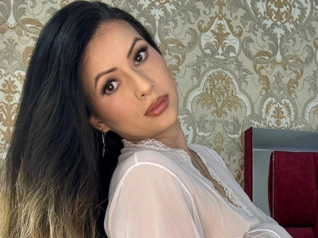 HannaRobertss has long black hair with blonde tips, brown eyes, and is wearing a sheer white top with lace. Her tits are small, and she's looking at the camera.