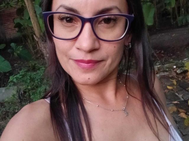 SolaraDubois has dark glasses, pink lipstick, and straight black hair. She's wearing a white tank top and a silver necklace with a small cross. Her tits are visible.