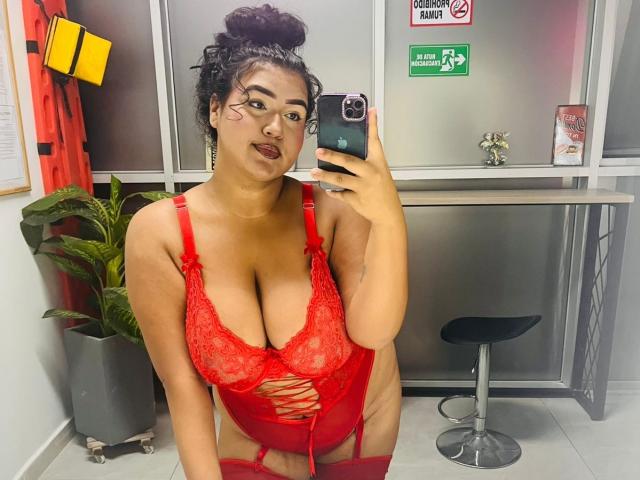 DojaAissha in red lace lingerie with big boobs, lace-up front, garter belt, and thigh straps, holding phone, dark curly hair in bun, pouty lips, mirror selfie.