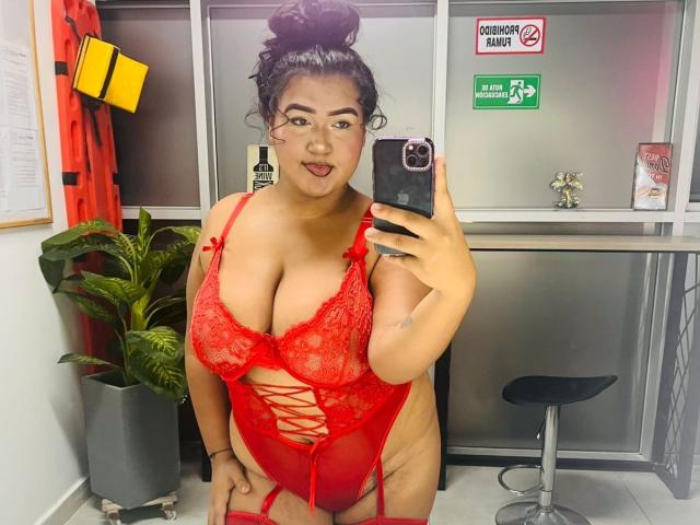 DojaAissha, curvy with large tits, wearing red lace lingerie, has her tongue out, taking a mirror selfie with her phone, black hair in a bun, in a room.