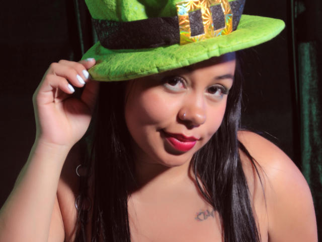 DojaAissha with large tits, black wavy hair, and a slight pout, looking at the camera, topless, in a bedroom setting. She has a cute, curvy, Latina face.