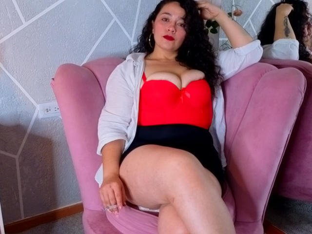 ArianaCherry with curly black hair, red strapless top showing her big boobs, black skirt, white shirt, pink chair, and red lipstick, sitting with one leg up.