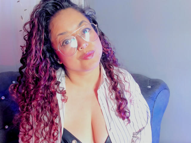SophiLarusso with curly red-brown hair, glasses, and pouty lips, wearing a striped shirt that reveals her big boobs and black bra. She's sitting on a blue tufted couch.