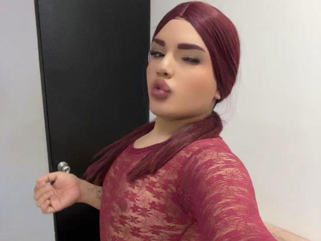 GlossyyBratz mit roten Haaren im Pferdeschwanz, voller Lippen, trägt eine durchsichtige rote Spitzenbluse, hält an einem Türgriff fest und zeigt ein Tattoo am linken Handgelenk..
