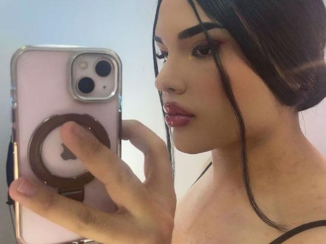 GlossyyBratz avec des lèvres roses brillantes, tenant un téléphone rose avec un support pour anneau, prenant un selfie, montrant son visage et ses cheveux noirs tressés sur le côté. Les seins ne sont pas visibles..