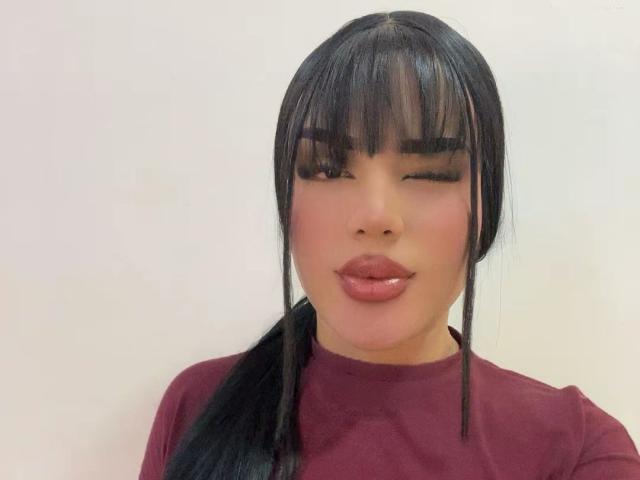 A GlossyyBratz tem cabelos pretos brilhantes em um rabo de cavalo, vestindo uma camisa vermelha, com lábios rosa cheios e um olho fechado em um piscar de olhos, parecendo fofa e sexy..