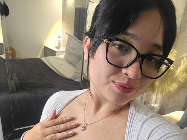 LunaBellaHotty with short black hair, wearing black glasses, white top, and gold necklace, showing pink lipstick, touching chest, and pink nails, smiling in selfie.