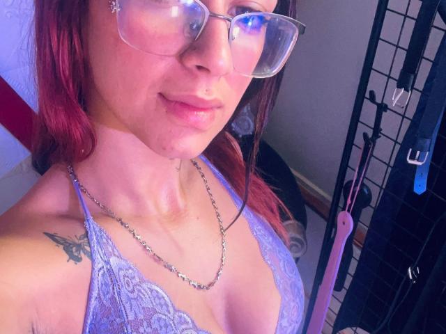 RoxanaSanchezz, con gli occhiali, i capelli rossi e un piercing al naso, indossa un reggiseno di pizzo blu, mostrando un tatuaggio sulla spalla sinistra e una collana d'argento, con un sex toy sullo sfondo..