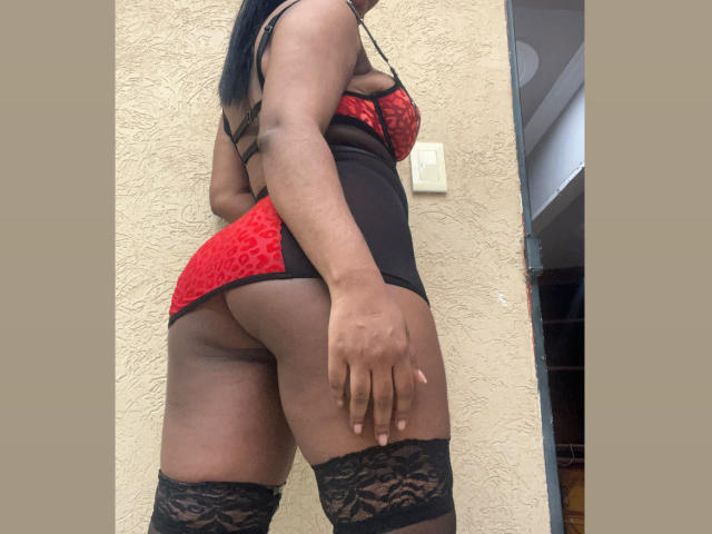 MelanyScoth in red and black lace lingerie, showing her ass, black thigh-high stockings, and a black strap bra. She's standing against a beige wall, looking back.