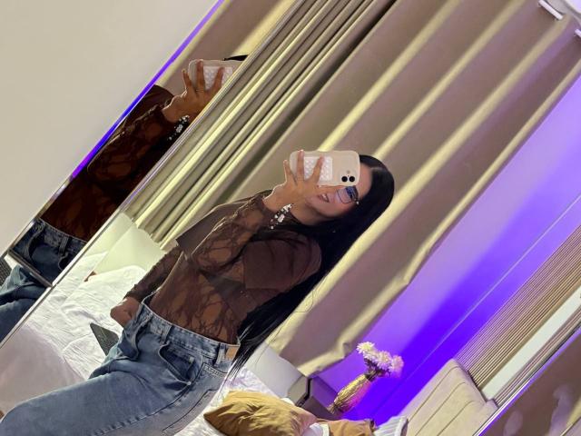 Antonella Preschot, com longos cabelos pretos, vestindo uma blusa marrom transparente e jeans azuis, tirando uma selfie no espelho, mostrando seus seios e nádegas, segurando um celular rosa..