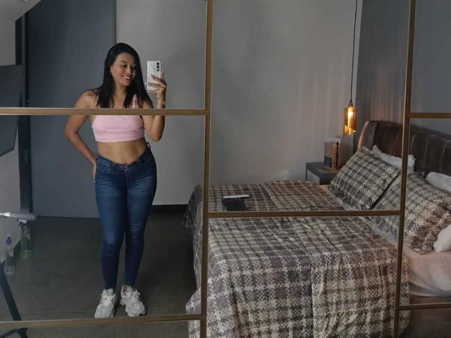 ZammySanz, con lunghi capelli neri, un top corto rosa che mostra il seno, jeans blu, scarpe da ginnastica bianche, in piedi davanti a uno specchio, che si fa un selfie in camera da letto con una coperta a quadri..