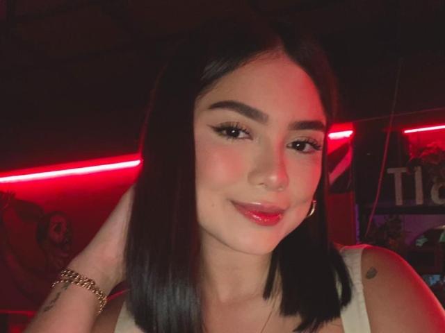 SkyBrownn with straight black hair, dark eyebrows, red lipstick, gold bracelet, and nose ring, smiling at the camera in red-lit environment. Her tits are slightly visible.