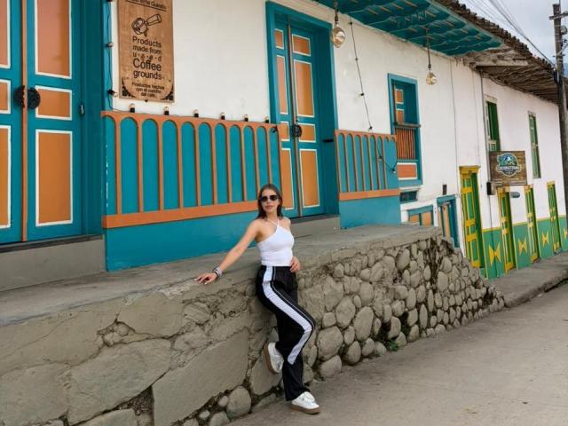 AnnaFer with medium tits, wearing a white tank top and black track pants with white stripes, leaning against a stone wall, standing in front of a colorful building.