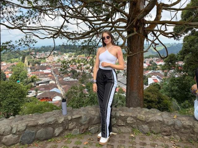 AnnaFer stands in black and white track pants, white top, and white sneakers, wearing sunglasses, with a tree behind her and a town below, showing her tits and ass.