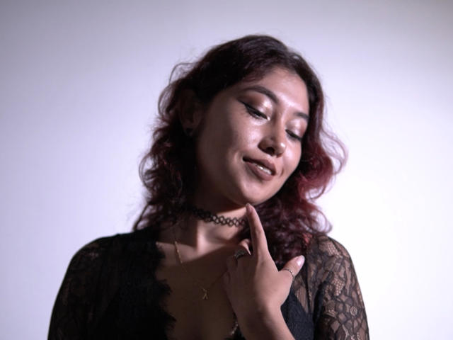 AliCipher has dark curly hair, a black choker, and a sheer black lace top, touching her neck with a subtle smile, showing her small tits and a nose ring.