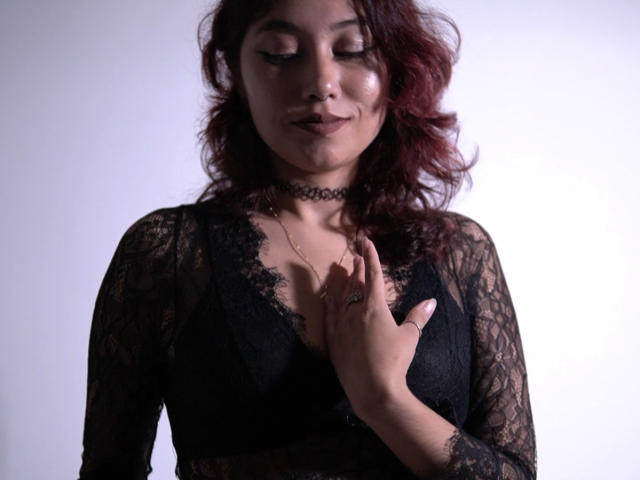 AliCipher with wavy red hair, black lace top, choker, and ring, hand on chest, eyes closed, smiling subtly. Boobs visible through lace. Soft light, gentle expression.