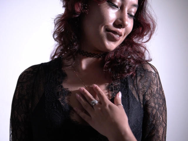 AliCipher with red curly hair, black lace top, choker, and ring, touching her chest, smiling, showing small tits, soft lighting, medium shot, realistic, sensual, confident.
