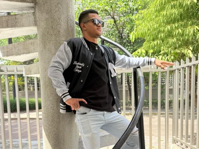 MaikolTwinkX leans against a spiral staircase, wearing a black and gray varsity jacket, black shirt, and light blue ripped jeans, with sunglasses on and a relaxed expression.