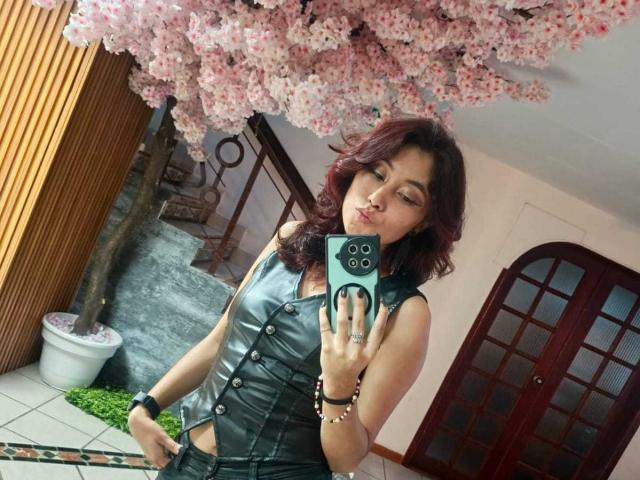 AliCipher with red hair, black leather sleeveless top, and bracelet, pouting at mirror, holding green phone, pink cherry blossom tree above, red door in background.
