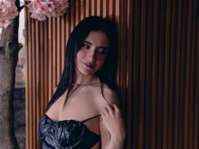 ValerieBanks with black leather strapless top, showing small tits, standing against wooden wall, holding strap with left hand, long black hair, pink cherry blossoms in background, subtle smile.