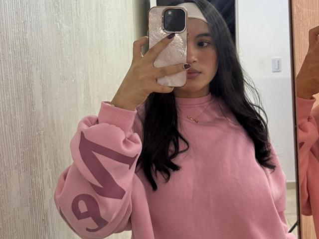 MiiaPalmer in a pink sweater with pink lettering, holding a pink phone, mirror selfie, long black hair, subtle makeup, pink nail polish, pink headband, calm expression.