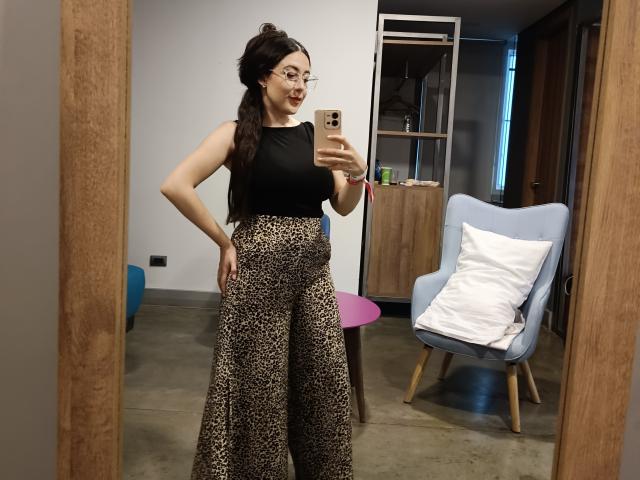 MabyLegrand with glasses, black sleeveless top, and leopard print pants, holding a phone, standing in front of a mirror, smiling, showing her ass.
