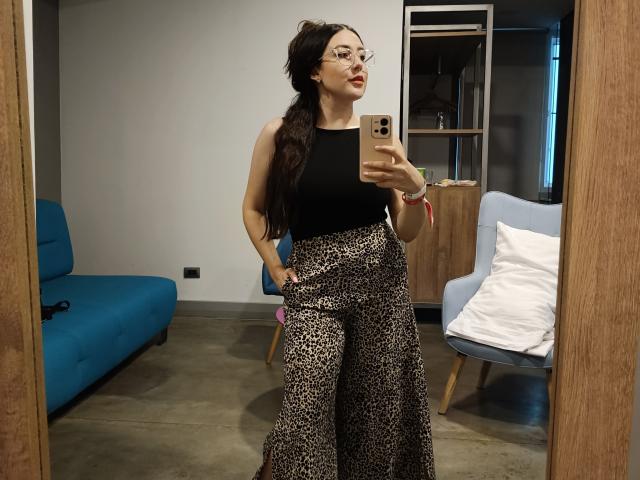 MabyLegrand with long brown hair, glasses, black sleeveless top, and leopard print pants, taking a mirror selfie, holding a gold phone, in a modern room.