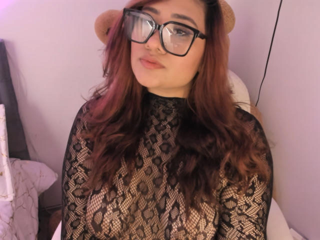 ElyRoss with glasses, wearing a black lace top that shows her tits, has long brown hair, and a cute face, sitting in a chair with bear ears on.