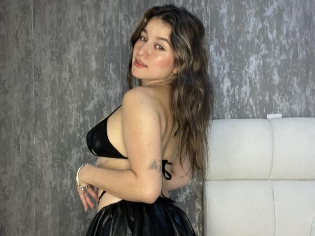 MilaGonz with wavy brown hair, black bikini top, and black skirt, showing her back, small tattoo on right arm, looking over her shoulder, white headboard in background.