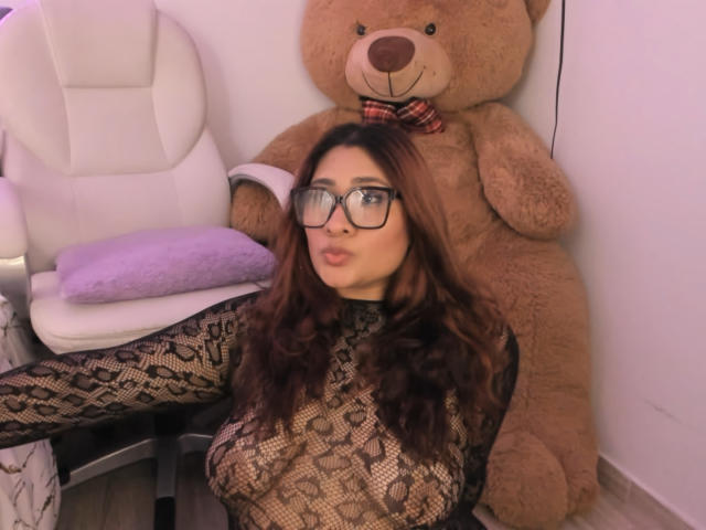 ElyRoss with glasses, long brown hair, big tits in a lace top, sitting in front of a giant teddy bear wearing a plaid bowtie, white chair, purple pillow.
