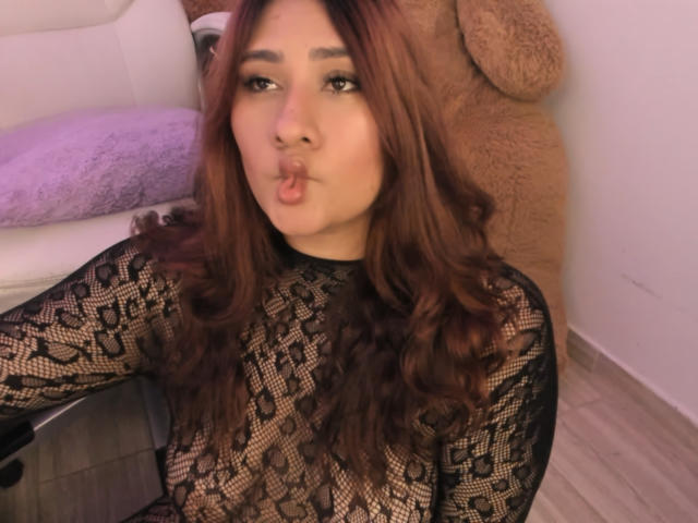 ElyRoss with long brown hair, pouty lips, wearing a black lace top, sitting in front of a giant brown teddy bear, looking into the camera.